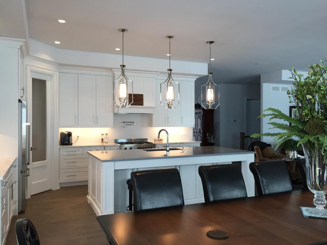 White kitchen cabinetry and island with lantern pendants, viewed across a wood dining table